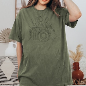 May include: A green t-shirt with a black line drawing of a camera with flowers on top.