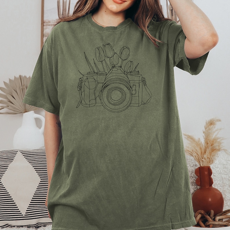 May include: A green t-shirt with a black line drawing of a camera with flowers on top.