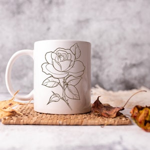 May include: White ceramic mug with a black line drawing of a rose on the front. The mug is sitting on a woven placemat.