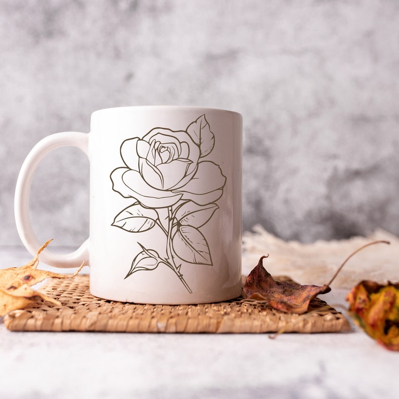 May include: White ceramic mug with a black line drawing of a rose on the front. The mug is sitting on a woven placemat.