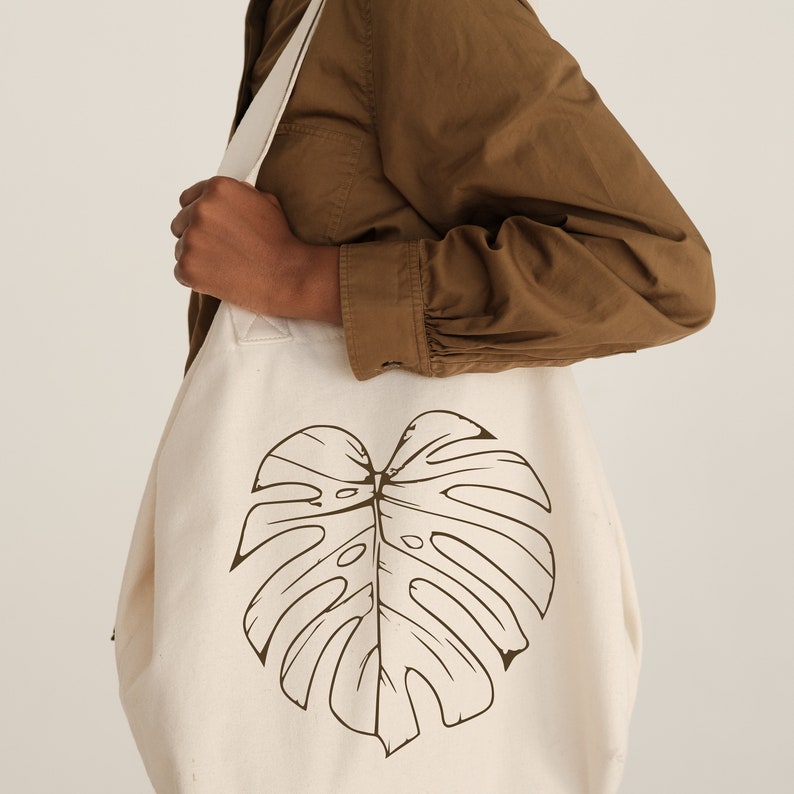 K&ouml;nnte beinhalten: Eine wei&szlig;e Canvas-Tragetasche mit einem braunen Monstera-Blatt-Design. Die Tasche hat einen braunen Riemen und wird von einer Person in einem braunen Hemd gehalten.