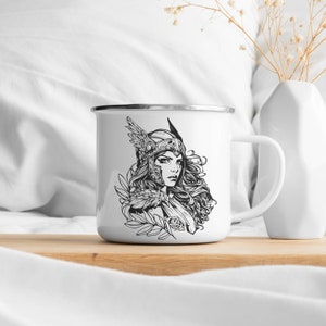 May include: White enamel mug with a black and white illustration of a woman wearing a helmet with wings. The woman has long hair and is looking to the right.