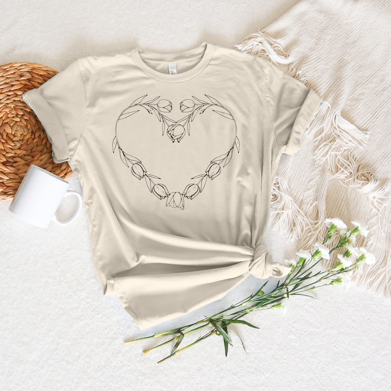 May include: A beige t-shirt with a black line drawing of a heart made of tulips.