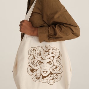 May include: A white canvas tote bag with a brown line drawing of Medusa, a woman with snakes for hair, on the front.