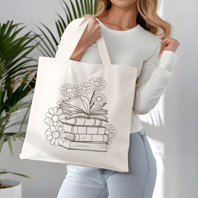 May include: A white canvas tote bag with a black and white line drawing of a stack of books with flowers on top. The bag is being held by a person.