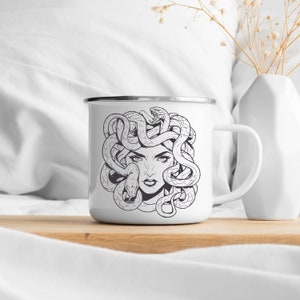 May include: White enamel mug with a black and white illustration of Medusa, a woman with snakes for hair.