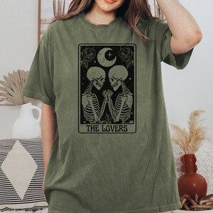 Może przedstawiać: Zielony T-shirt z czarno-białą ilustracją karty tarota Miłosnych, przedstawiającą dwa szkielety trzymające się za ręce. Karta ma p&oacute;łksiężyc i gwiazdy nad szkieletami oraz r&oacute;że po obu stronach. Tekst "THE LOVERS" jest nadrukowany pod szkieletami.
