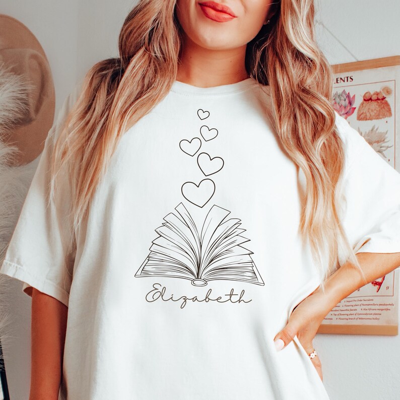 May include: White t-shirt with a black line drawing of an open book with hearts floating above it. The text "Elizabeth" is written below the book.