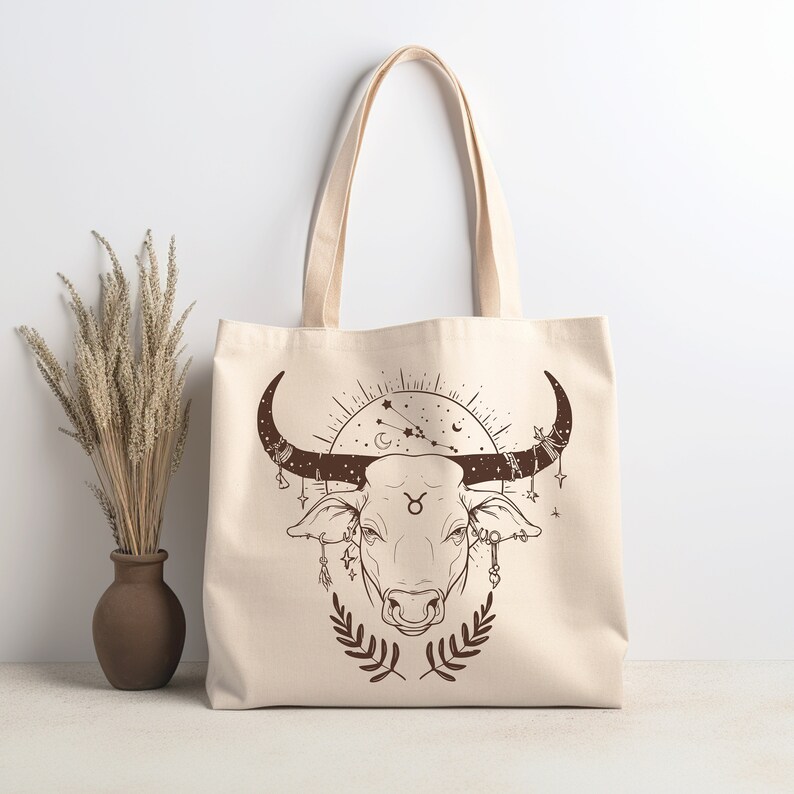May include: A beige canvas tote bag with a brown illustration of a bull's head with horns, surrounded by stars and a crescent moon. The bull's head has a Taurus symbol on its forehead.