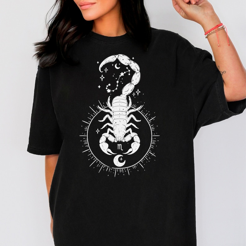 May include: Black t-shirt with a white graphic of a scorpion with a crescent moon and stars. The scorpion is surrounded by a circle with a crescent moon and the letter "m" in the center.
