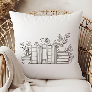 May include: A white throw pillow with a black and white illustration of a stack of books, a bird, a cup, and plants. The illustration is centered on the pillow.