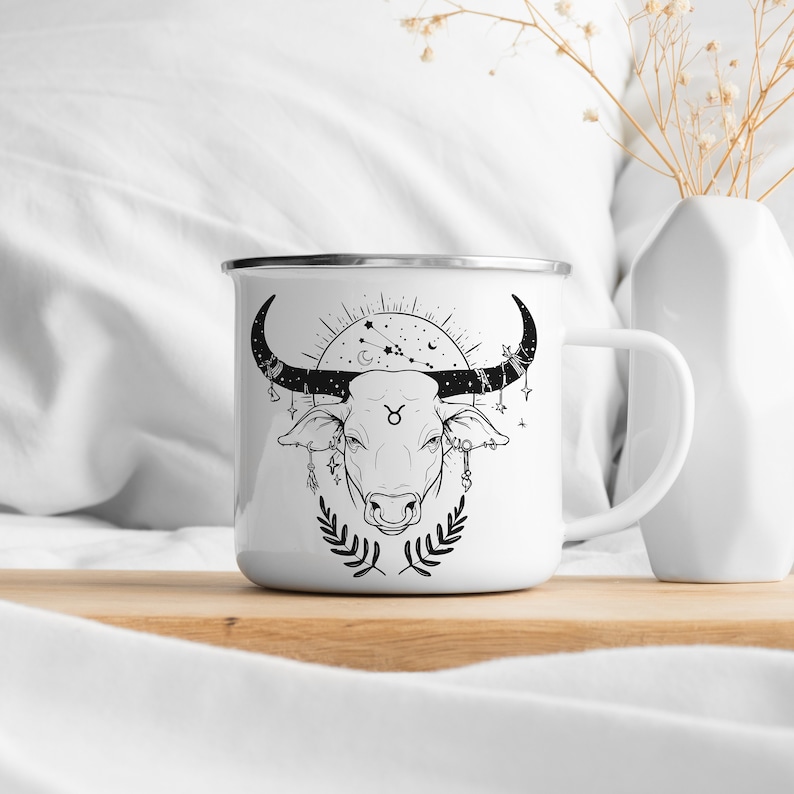 May include: White enamel mug with a black and white illustration of a bull's head with a Taurus zodiac symbol and celestial details. The mug has a silver rim and handle.