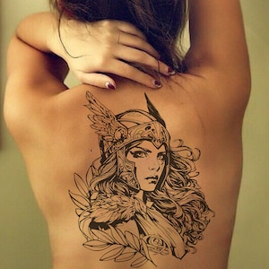 May include: A black and white line art tattoo design of a woman wearing a winged helmet and armor. The design features intricate details and a flowing hairstyle.