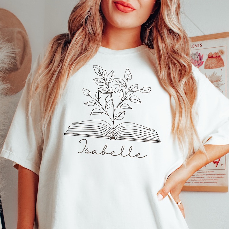 May include: White t-shirt with a black line drawing of an open book with a plant growing out of it. The text "Isabelle" is written below the book.