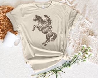 Op de afbeelding: Een beige t-shirt met een vintage illustratie van een cowboy die een opspringend paard berijdt. De illustratie is in tinten bruin en zwart.