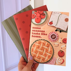 Op de afbeelding: Drie rechthoekige wenskaarten. De voorste kaart bevat een illustratie van een appeltaart, een kopje koffie, appelschijfjes en een open receptenboek met de tekst 'some seasons taste like home.' De andere twee kaarten zijn rood met stippen en olijfgroen met strepen.