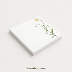 May include: A stack of square white notepads with a watercolor daisy design. The daisy has white petals and a yellow center, with green stems and leaves. The bottom left corner of the image has the text "lavendergreen. studio."