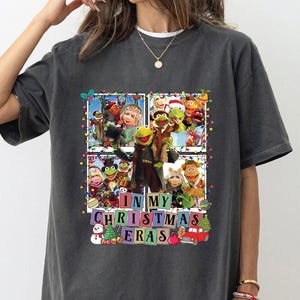 May include: Dark gray t-shirt featuring a colorful graphic of Muppet characters in a Christmas theme. The design includes the text "IN MY CHRISTMAS ERAS" in a block letter style, with festive elements like a snowman and a Christmas tree.