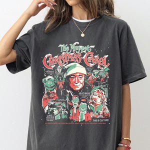 May include: Dark gray t-shirt featuring a graphic from "The Muppet Christmas Carol." The design includes characters from the movie, with red and green accents. The text "The Muppet Christmas Carol" is prominently displayed.