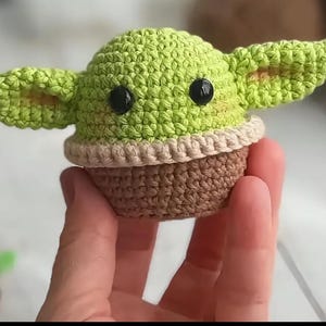 May include: A crocheted figure of a character with large ears, black eyes, and a light green head. The figure has a tan and brown body. The figure is held in a person's hand.