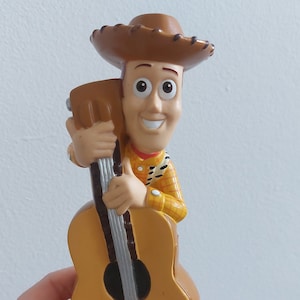 May include: A Woody figurine from the Toy Story franchise, holding a guitar. The figure is wearing a brown cowboy hat, a yellow shirt with a red bandana, and a sheriff's badge. The guitar is a light brown color.