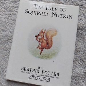 Peut inclure: Couverture du livre "L'Histoire d'Écureuil Nutkin" illustrant un écureuil roux à la queue touffue tenant une noix. Le livre est de Beatrix Potter et est l'édition originale et autorisée publiée par F. Warne & Co.
