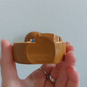 May include: A brown Disney/Pixar figurine with the text "&copy;Disney/Pixar China" on the base. The figurine is held in a hand. The figurine has a light blue and tan color scheme.