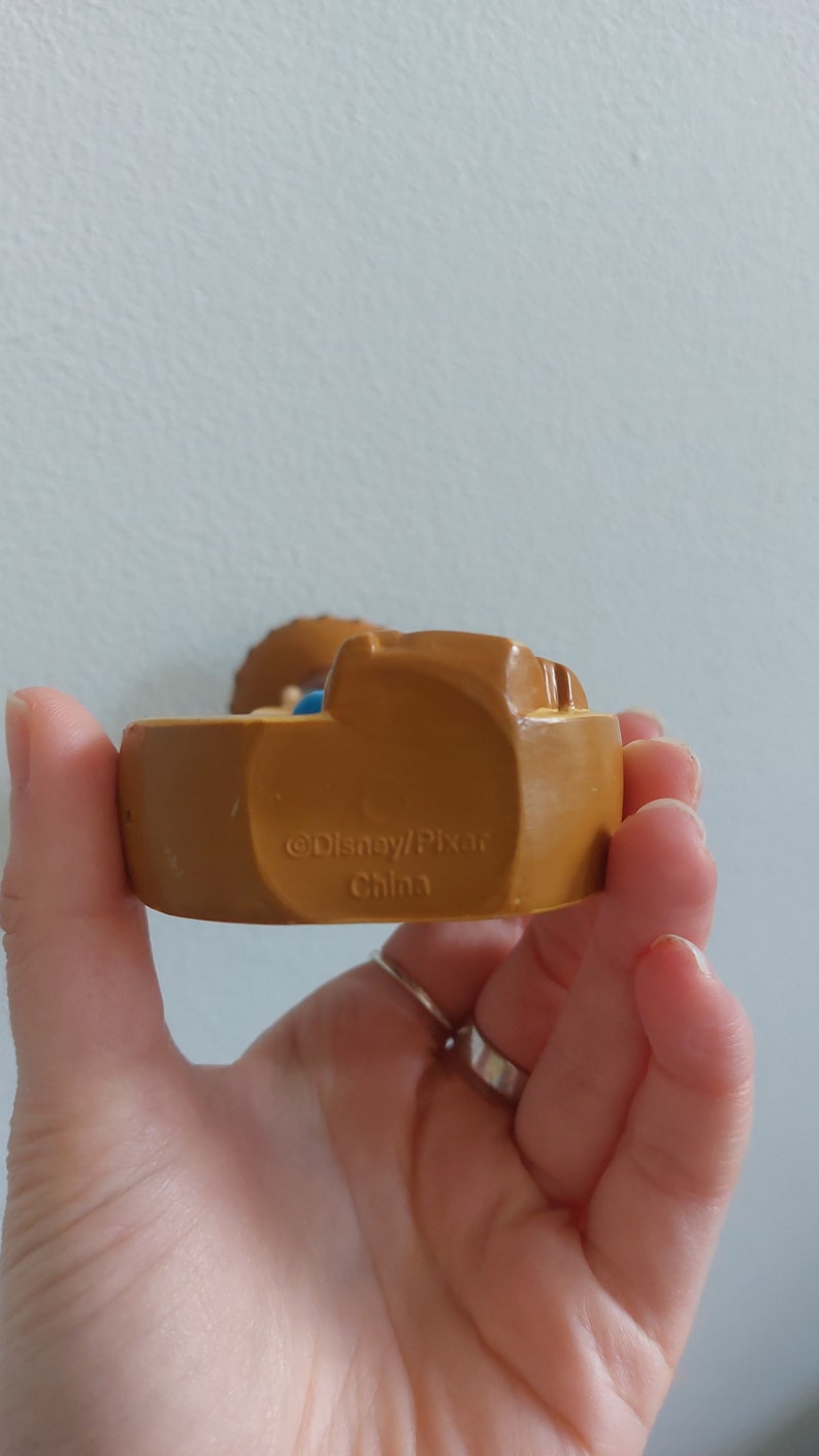 May include: A brown Disney/Pixar figurine with the text "&copy;Disney/Pixar China" on the base. The figurine is held in a hand. The figurine has a light blue and tan color scheme.