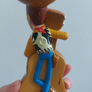 May include: A plastic toy figurine of Woody from Toy Story. Woody is wearing a brown cowboy hat, a yellow plaid shirt, a black and white vest, blue pants, and brown boots. The figurine is mounted on a brown guitar.