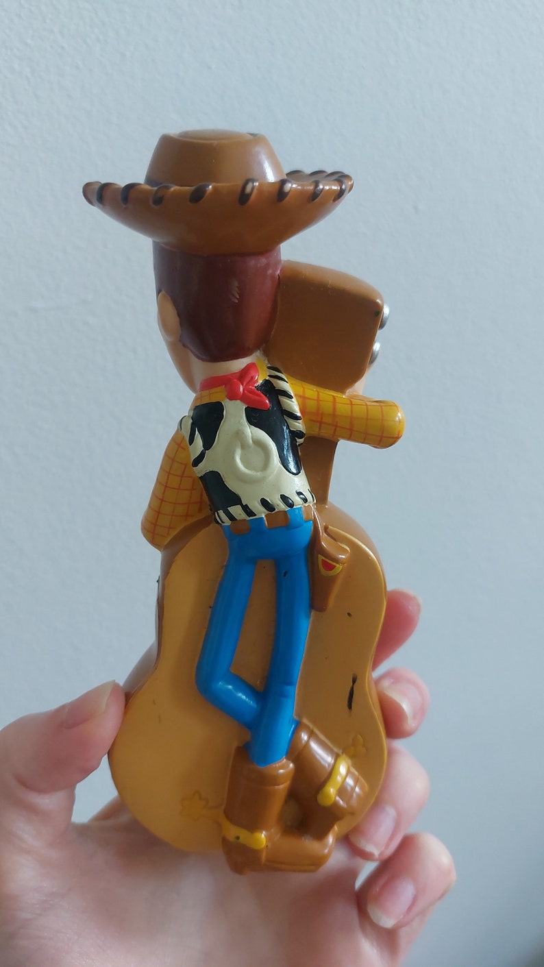May include: A plastic toy figurine of Woody from Toy Story. Woody is wearing a brown cowboy hat, a yellow plaid shirt, a black and white vest, blue pants, and brown boots. The figurine is mounted on a brown guitar.