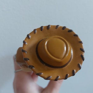 May include: A close-up of a tan cowboy hat with a brown brim and stitching detail. The hat has a rounded crown and a wide brim, with a dark brown stitched border. The hat is a toy or collectible item.