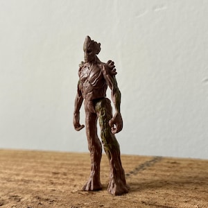May include: A brown and green plastic action figure of a tree-like humanoid character. The figure has a detailed, textured surface and is posed standing with its arms at its sides.