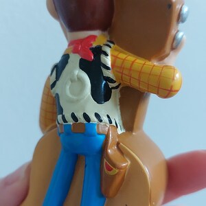 May include: A figurine of the Toy Story character Woody, seen from the back. Woody wears a brown cowboy hat, a white vest with a black cowhide pattern, blue jeans, and a red neckerchief. He has a brown holster.