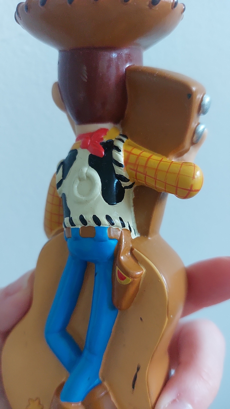 May include: A figurine of the Toy Story character Woody, seen from the back. Woody wears a brown cowboy hat, a white vest with a black cowhide pattern, blue jeans, and a red neckerchief. He has a brown holster.