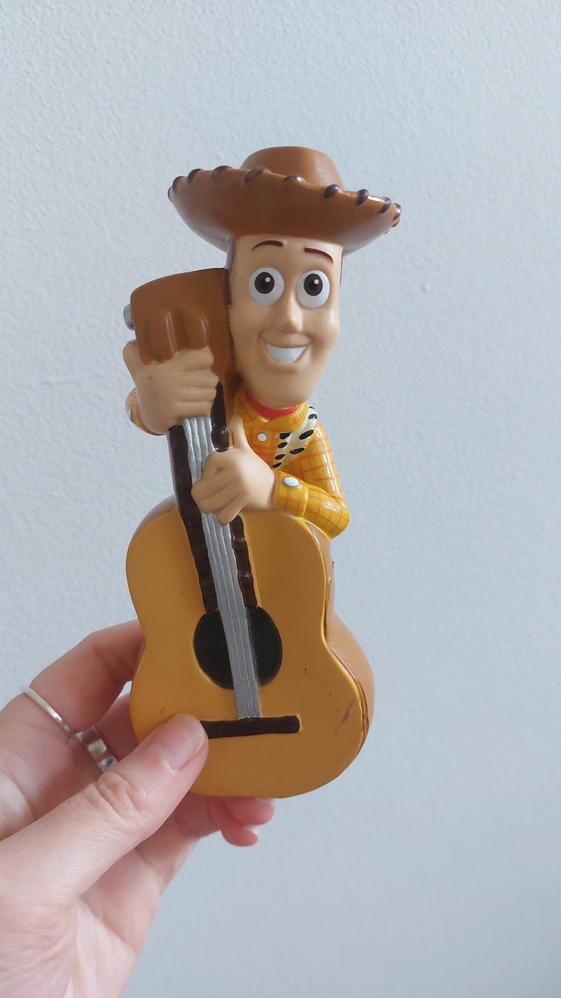 May include: A Woody figurine from the Toy Story franchise, holding a guitar. The figure is wearing a brown cowboy hat, a yellow shirt with a red bandana, and a sheriff's badge. The guitar is a light brown color.