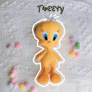 May include: A crocheted Tweety Bird plush toy with yellow feathers, blue eyes, and a black beak. The toy is sitting on a white crocheted surface.