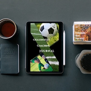 Puede incluir: Una tableta negra con un lápiz blanco a su lado. La pantalla de la tableta muestra una portada de diario digital con el texto "GRASSROOTS COACHING JOURNAL" e imágenes de jugadores de fútbol y un balón de fútbol. La tableta está sobre una superficie gris con una libreta negra, un bolígrafo, una taza de café, una pequeña caja de café y un objeto hexagonal.
