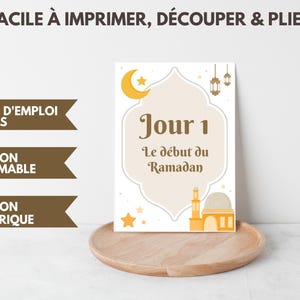 以下が含まれることがあります： 「Jour 1 Le d&eacute;but du Ramadan」のテキストが入った、白とベージュのラマダンをテーマにしたプリント。三日月、星、モスクのイラストが特徴です。「FACILE &Agrave; IMPRIMER, D&Eacute;COUPER & PLIER」の追加テキストが含まれています。
