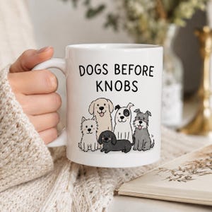 May include: White ceramic mug featuring the text "DOGS BEFORE KNOBS" and a cartoon illustration of six different dog breeds. The mug is held by a person wearing a cream-coloured sweater.