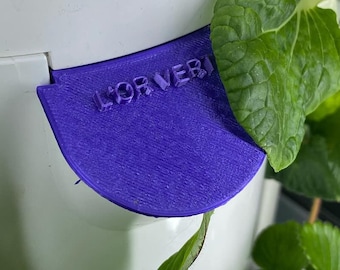 May include: A white plastic vertical garden tower with a purple plastic water reservoir lid. The lid has the text "FOR VERMI" printed on it. The tower is filled with green leafy plants.