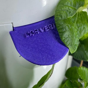 Peut inclure: Une tour de jardin verticale en plastique blanc avec un couvercle de réservoir d'eau en plastique violet. Le couvercle porte l'inscription "FOR VERMI". La tour est remplie de plantes vertes feuillues.