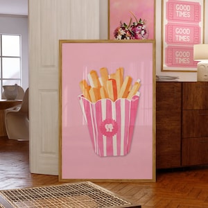 May include: A framed pink print featuring a watercolor illustration of french fries in a pink and white striped container with the Barbie logo. The print is displayed with other framed art pieces in a room with wooden floors.