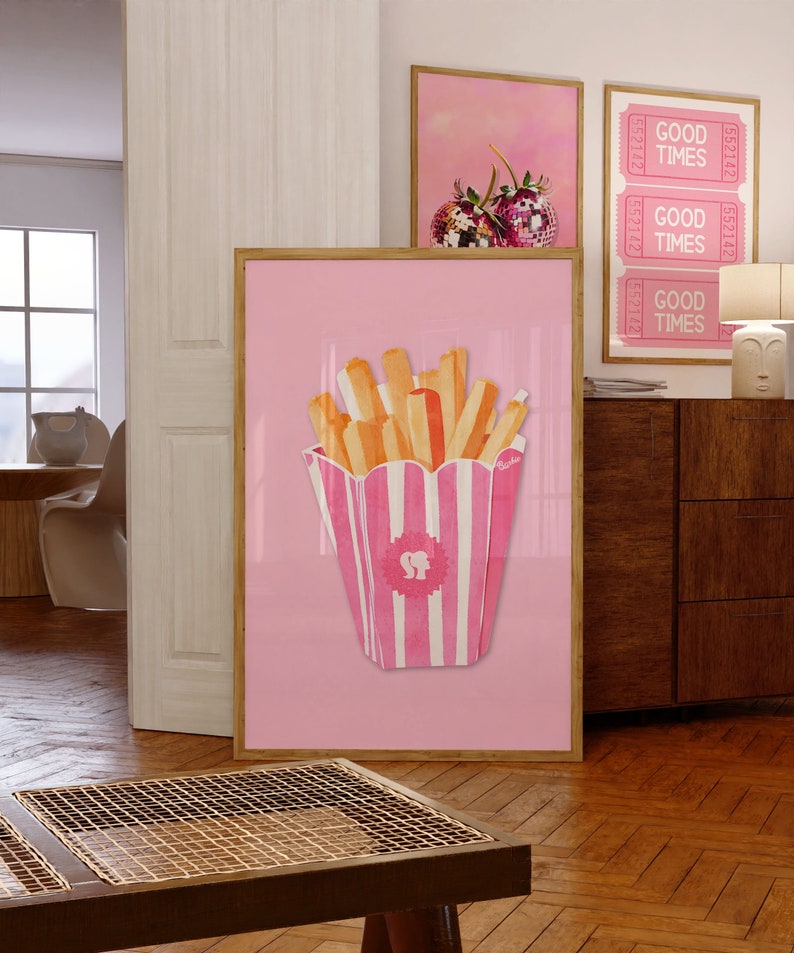 May include: A framed pink print featuring a watercolor illustration of french fries in a pink and white striped container with the Barbie logo. The print is displayed with other framed art pieces in a room with wooden floors.