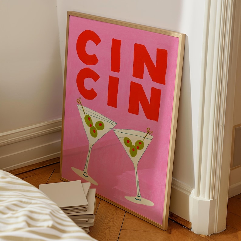 Op de afbeelding: Een ingelijste roze kunstprint met het woord "CIN CIN" in grote rode letters. Twee martini glazen met olijven zijn afgebeeld onder de tekst. De print heeft een gouden lijst en leunt tegen een muur.
