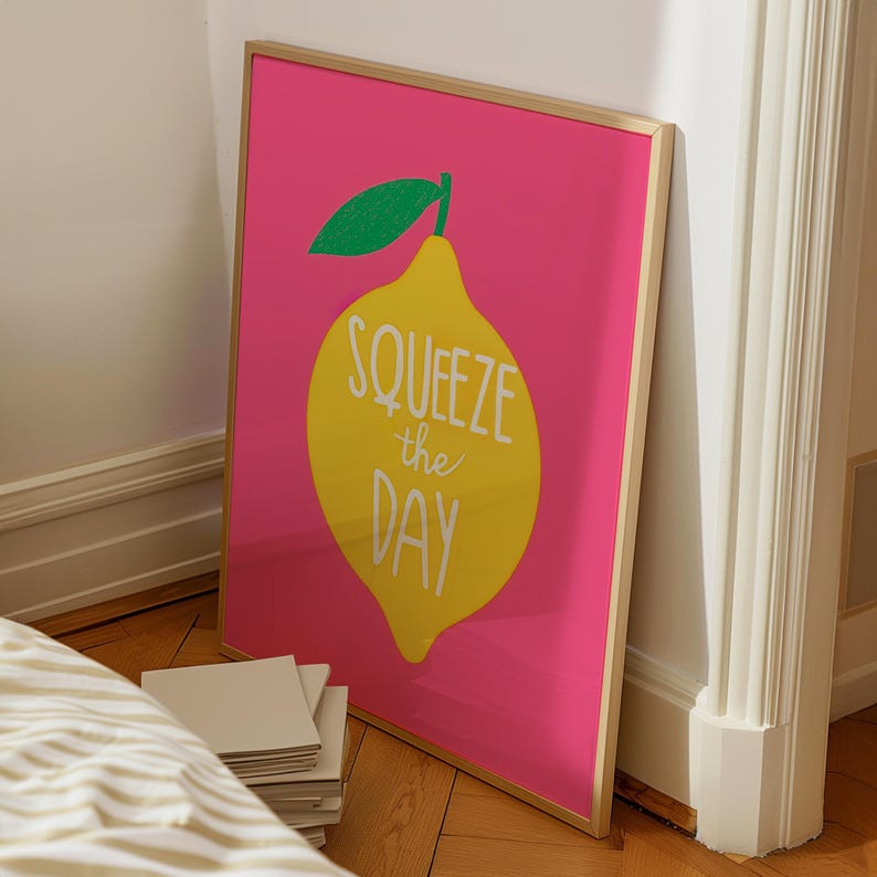 Op de afbeelding: Een ingelijste print met een gele citroen en een groen blad op een felroze achtergrond. De woorden "Squeeze the Day" staan in het wit op de citroen. Het goudkleurige frame leunt tegen een muur, wat een vrolijke uitstraling cre&euml;ert.