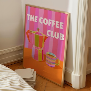 Op de afbeelding: Een ingelijste poster met de tekst "THE COFFEE CLUB" en een kleurrijke illustratie van een koffiezetapparaat en een mok. De achtergrond heeft verticale strepen in roze, oranje en paarse tinten. Het koffiezetapparaat en de mok zijn ook felgeleurd.