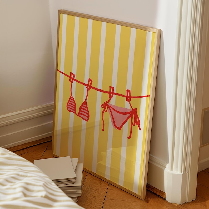 Op de afbeelding: Ingelijste kunst met een geel en wit gestreepte achtergrond. Een rode lijn met wasknijpers houdt een rode en roze bikini vast. Het kunstwerk leunt tegen een witte muur en een houten vloer.