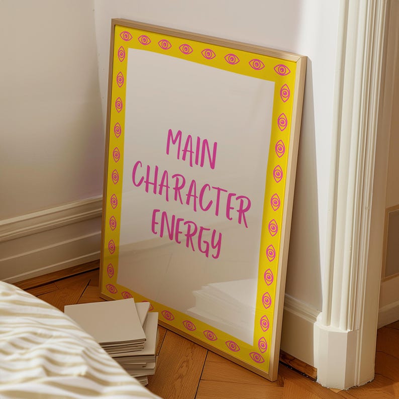 Op de afbeelding: Een ingelijste kunstprint met de woorden "MAIN CHARACTER ENERGY" in roze. De lijst heeft een gele rand met herhaalde roze oogontwerpen. De print leunt tegen een witte muur en een houten vloer.