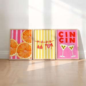 May include: Three framed art prints. The first features orange slices on a pink and white striped background. The second shows a red bikini hanging on a yellow and white striped background. The third has two martini glasses and the text "CIN CIN" on a pink background.