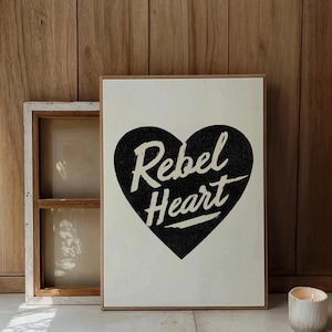 May include: A framed canvas print with the words "Rebel Heart" in white script inside a black heart shape. The background is off-white. A wooden window frame and a lit candle are in the background.